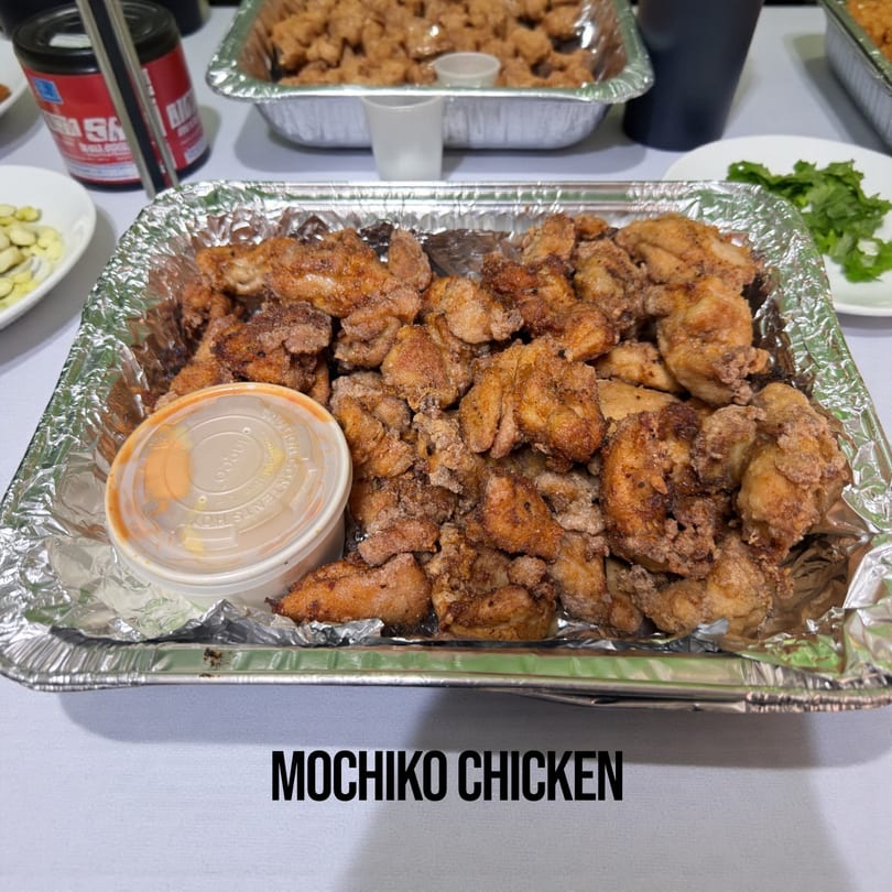 A foil pan filled with golden-brown mochiko chicken pieces and a small container of dipping sauce.